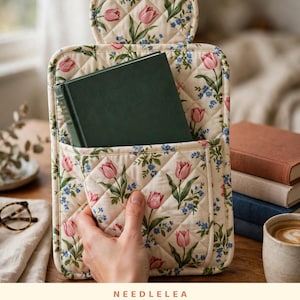 May include: A quilted book sleeve featuring a cream background with a floral pattern of pink tulips and small blue flowers. The sleeve has a tulip-shaped top flap and a front pocket containing a dark green hardcover book.