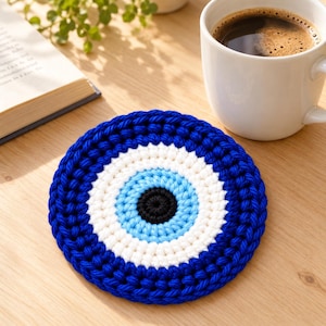 Peut inclure: Un dessous de verre circulaire au crochet arborant un motif d'œil concentrique. Le design se compose d'un centre noir uni, suivi d'anneaux bleu clair, blancs et d'une bordure extérieure épaisse en fil bleu foncé, formant un accessoire textile coloré.