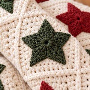 Può includere: Una coperta all'uncinetto color crema caratterizzata da un motivo a rombi ripetuto e da stelle a cinque punte che si alternano tra il verde scuro e il rosso intenso. Il tessuto è realizzato con punti visibili che creano una superficie.