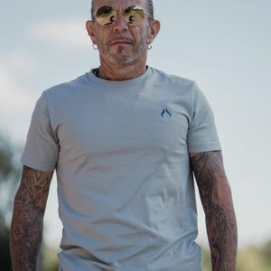 Könnte beinhalten: Ein hellgraues Kurzarm-T-Shirt mit einem kleinen, dunkelblauen, flammenförmigen Logo auf der linken Brust. Das T-Shirt hat einen Rundhalsausschnitt und eine schlichte, einfarbige Stoffkonstruktion, die einen sauberen und einfachen Look für den Alltag bietet.