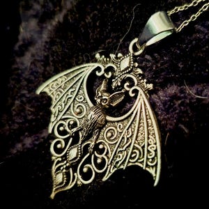 Puede incluir: Collar con colgante de murciélago plateado. El colgante presenta un diseño detallado de murciélago con alas extendidas, intrincados adornos y una pequeña figura de murciélago. El collar tiene una cadena delicada y un cierre seguro, perfecto para un toque gótico.