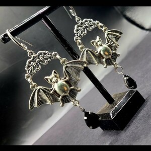 May include: A pair of silver bat earrings with black teardrop-shaped stones hanging from the bottom. The bats are detailed with wings and have a 3D effect.