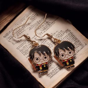 May include: A pair of gold-tone dangle earrings featuring a cartoon character with black hair, glasses, and a red and yellow scarf. The earrings are set against a backdrop of aged paper.