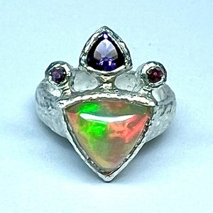 May include: A silver ring with a triangular opal gemstone set in the center. The ring is adorned with three smaller gemstones: a purple amethyst and two red rubies.