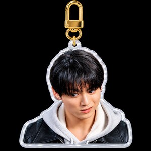 May include: A clear acrylic keychain featuring a portrait of a person with dark hair, wearing a gray hooded sweatshirt under a black jacket. The keychain includes a gold-colored metal lobster clasp at the top for attachment.