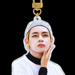 May include: An acrylic keychain featuring a portrait of a person with blonde hair wearing a grey beanie, a black mock neck shirt, and a white t-shirt, with white skincare cream applied to their face. The keychain has a gold-toned metal clasp.