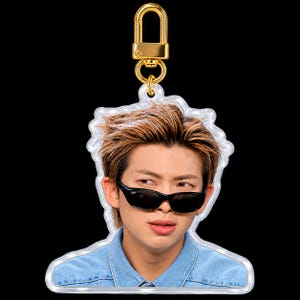May include: An acrylic keychain featuring a portrait of a person with brown hair wearing black sunglasses and a blue denim shirt. The keychain has a gold-colored metal lobster clasp attached to the top of the clear acrylic cutout.