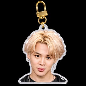 May include: A clear acrylic keychain featuring a portrait of a person with light blonde hair, wearing silver hoop earrings and a silver chain necklace. The keychain is attached to a gold-colored metal lobster clasp hardware.
