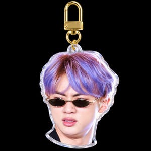 May include: An acrylic keychain featuring a portrait of a person with purple-toned hair wearing small, oval-shaped dark sunglasses. The charm is attached to a gold-colored metal lobster clasp and swivel hook hardware.