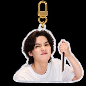 May include: An acrylic keychain featuring a person with dark hair wearing a white t-shirt, making a determined facial expression while holding a pair of brown chopsticks. The charm is attached to a shiny gold-toned metal lobster clasp.