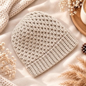 May include: A beige crochet beanie featuring a textured star-stitch pattern on the crown and a ribbed brim. The hat is crafted from yarn and displays a consistent light tan color throughout, showcasing the intricate hand-stitched details.