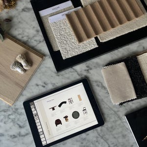 Op de afbeelding: Een verzameling interieurontwerpmaterialen met stoffenstalen in crème, zwart en beige, een licht houten paneel met twee kleine garenbundels, een zwarte lade met getextureerde textielmonsters en een tablet met een meubelcatalogus.