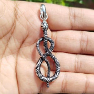 Può includere: Un ciondolo in metallo color argento a forma di serpente avvolto che forma un simbolo di infinito. La superficie presenta un motivo a scaglie dettagliato e testurizzato su tutto il corpo, con un occhiello liscio e lucido sulla parte superiore.