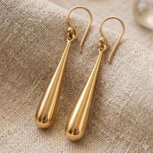 May include: A pair of gold-toned drop earrings featuring a smooth, elongated teardrop shape. Each earring is suspended from a curved wire hook, showcasing a polished, reflective metallic surface that tapers to a rounded point at the bottom end.