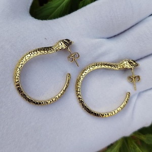 Peut inclure: Une paire de boucles d'oreilles créoles dorées en forme de serpent. Chaque boucle présente une surface texturée rappelant des écailles le long du corps incurvé et une tête de serpent détaillée sur le dessus, avec un fermoir à tige.