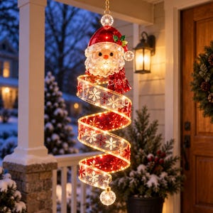 Op de afbeelding: Een hangende kerstversiering met een kerstmanhoofd met een rode muts en witte baard, bevestigd aan een spiraalvormig, verlicht rood en goud lint versierd met witte sneeuwvlokpatronen, eindigend in een kleine heldere lichtgevende bol.