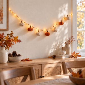 May include: A decorative garland featuring small crocheted pumpkins in shades of orange, white, and beige, interspersed with warm glowing fairy lights and wooden beads, suspended horizontally against a plain wall above a wooden surface.