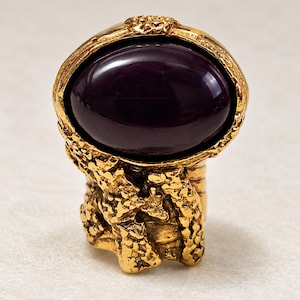 Op de afbeelding: Een goudkleurige ring met een grote, gladde, ovaalvormige donkerpaarse steen in een textuurrijke, ruwe metalen band. Het metalen oppervlak heeft een poreus, onregelmatig uiterlijk dat contrasteert met het gepolijste, reflecterende oppervlak van de steen.