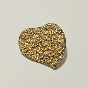 Puede incluir: Un broche en forma de corazón de tono dorado con una superficie texturizada y granulada. El objeto tiene un borde liso y elevado que perfila la forma del corazón, con un acabado denso, irregular y reflectante en toda la cara frontal.