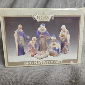 May include: A rectangular box for a Crown Accents 6PC Nativity Set. The packaging features an illustration of six porcelain-style figurines in blue, purple, and cream robes, including a central infant in a manger, all set against a dark background.