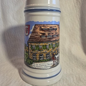 May include: A white ceramic beer stein featuring a colorful illustration of a stone building with green shutters and a horse-drawn carriage. The text reads 11. Int. Wandertag 1990 Wfr. Heubsch u. Umgebung. The stein has blue trim at the top and base.
