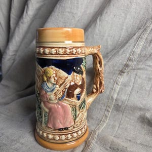 May include: A ceramic beer stein featuring a relief scene of a person playing a stringed instrument and another person sitting at a table. The stein has a tan rim and base, a dark blue background, and a handle shaped like a textured tree branch.