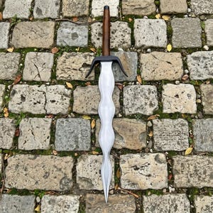 May include: A sword with a wavy, double-edged steel blade, a black metal crossguard, and a cylindrical dark brown wooden handle. The blade features a distinct undulating design that tapers to a sharp point, contrasting with the dark handle.