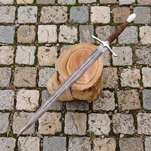 May include: A long sword with a patterned Damascus steel blade, a silver crossguard, and a brown wooden handle featuring intricate carvings. The pommel is silver and rounded, and the blade displays a distinct wavy, layered metal texture.