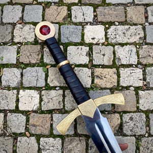 May include: A sword with a polished steel blade, a gold-colored crossguard featuring intricate etched patterns, and a black leather-wrapped grip. The pommel is a circular gold-colored disc set with a large, faceted, deep red gemstone.