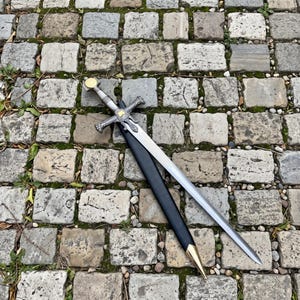 Op de afbeelding: Een zilverkleurig metalen zwaard met een puntig lemmet, een kruisvormige pareerstang met gedetailleerde gravures en een ronde, goudkleurige pommel. Het zwaard wordt vergezeld door een zwarte schede met een goudkleurige metalen punt.