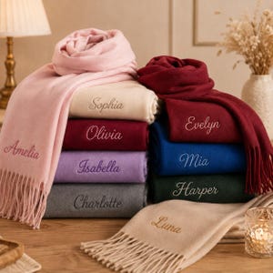 May include: A stack of soft, folded scarves in various colours including light pink, cream, maroon, lavender, grey, dark blue, and dark green. Each scarf features embroidered names: Amelia, Sophia, Olivia, Isabella, Charlotte, Evelyn, Mia, Harper, and Luna.