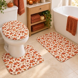 Op de afbeelding: Een badkamerset bestaande uit een toiletbrilhoes, een U-vormig contourkleed en een rechthoekige badmat, allemaal voorzien van een herhalend patroon van kleine witte spookjes in oranje dekens, oranje pompoenen en bruine herfstbladeren.