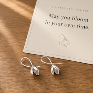 May include: A pair of silver-toned drop earrings shaped like snowdrop flowers, accompanied by a cream-colored card featuring the text A LITTLE GIFT FOR YOU May you bloom in your own time and a line drawing of a single snowdrop flower.