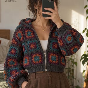 Peut inclure: Une veste à capuche zippée composée de carrés granny au crochet dans des tons gris foncé, bleu sarcelle et orange brûlé. Le vêtement présente une fermeture éclair en métal argenté, des poignets et un ourlet côtelés. Logo Farazana visible.