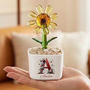 May include: A glass sunflower with yellow petals and a green stem, planted in a small white square ceramic pot filled with light-colored pebbles. The pot features a large red letter A decorated with floral patterns and the name Amelia below it.