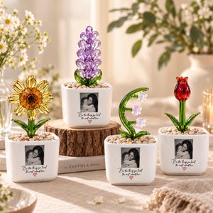 May include: Four white square ceramic planters, each containing a crystal flower and small stones. The planters feature a black and white photo of a person holding a child, with the text "Be the things you loved the most about her." and a red heart.