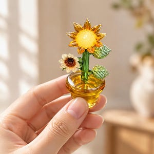 Può includere: Una statuetta in miniatura di girasole in vetro in un piccolo vaso color ambra. La scultura presenta un grande girasole giallo con un centro strutturato, un fiore bianco più piccolo e due foglie di vetro verde su uno stelo verde.