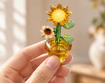 May include: A miniature glass sunflower figurine in a small amber-colored pot. The sculpture features a large yellow sunflower with a textured center, a smaller white flower, and two green glass leaves attached to a green stem.