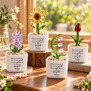 May include: Four glass flower sculptures in white square pots filled with small stones. Each pot features the text: May you be proud of the work that you do the person you are and the difference you make, Evelyn, 11.22.26, with a red heart.