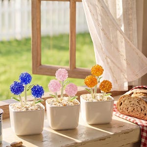 May include: Three square white ceramic pots each containing a small artificial plant with three round, textured flower heads. One plant has blue flowers, one has light pink flowers, and one has orange flowers, all with green stems.