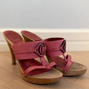 Op de afbeelding: Een paar roze leren instapsandalen met houten hakken en plateaus. De sandalen hebben een brede band over de wreef versierd met een rond zwart logo-embleem, dunne teenbandjes en kleine metalen studs die het leer aan het hout bevestigen.