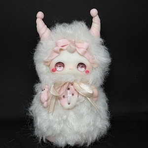 Peut inclure: Une créature en peluche recouverte de longs poils blancs avec deux cornes rose pâle sur la tête. Elle présente de grands yeux roses étincelants, un petit nœud rose sur le front et un ruban beige noué autour d'un gros bouton blanc sur la poitrine.