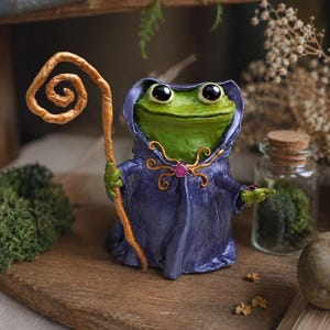 Könnte beinhalten: Eine grüne Froschfigur in einer dunkelvioletten Kapuzenrobe mit einer rosa Edelsteinbrosche. Der Frosch hält einen hohen, hellbraunen Holzstab mit spiralförmigem Ende in der einen Hand und einen kleinen dunklen Gegenstand in der anderen, mit großen schwarzen Augen.