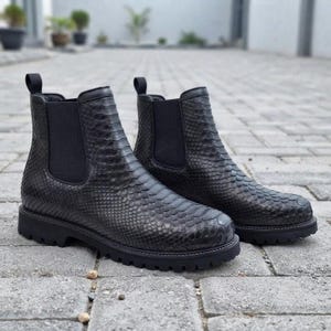 Op de afbeelding: Een paar zwarte enkellaarzen met een textuur van slangenleer, elastische zijpanelen en een dikke profielzool. Deze instaplaarzen hebben een ronde neus en een treklipje aan de achterkant van de schacht, uitgevoerd in een donkere, geschubde afwerking.