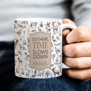 May include: A white ceramic mug featuring a repeating brown ink pattern of various wildlife, including deer, foxes, fish, and birds. A central tan rectangular label displays the text "OUT HERE, TIME SLOWS DOWN" followed by the name "MICHAEL".