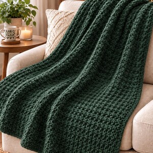 May include: A dark forest green chunky knit throw blanket with a textured, raised stitch pattern. The blanket is draped over a light beige upholstered sofa, showcasing the thick yarn construction and the deep, solid color of the textile.