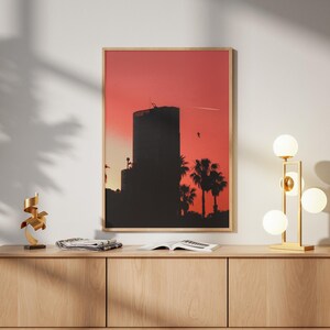 May include: A rectangular wall art print in a light wood frame featuring a silhouette of a tall building and palm trees against a vibrant red and orange sunset sky. The artwork is displayed above a long, light-toned wooden media console cabinet.