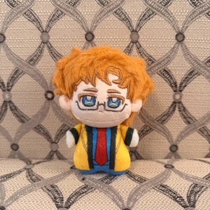 May include: A small plush doll with orange hair, blue embroidered eyes, and glasses. The doll wears a yellow jacket with a red tie and blue accents. The figure has a rounded head and a compact body, featuring detailed stitching on the face and clothing.