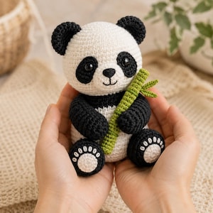 Könnte beinhalten: Ein kleines, handgefertigtes Häkel-Pandabärchen aus weißem und schwarzem Garn. Der Panda hat runde schwarze Ohren, schwarze Augenpartien sowie eine gestickte Nase und einen Mund. Er hält einen kleinen, strukturierten grünen Bambuszweig.