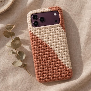Op de afbeelding: Een rechthoekig smartphonehoesje met een textuurrijke, handgehaakte hoes met een diagonaal kleurblokontwerp. Het hoesje is verdeeld in twee secties, een in romig gebroken wit garen en de andere in warm, gedempt terracotta oranje garen.